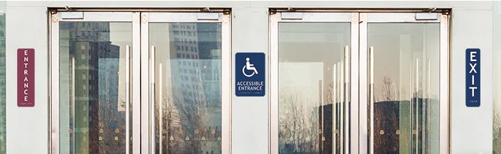 An Entrance Sign, Exit Sign, And Accessible Entrance Sign Posted Outside Of A Building With The Corresponding Braille Below