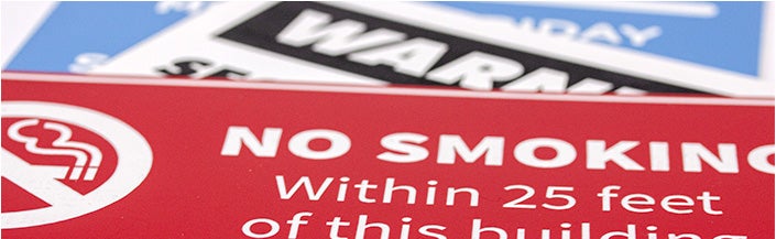 A red NO SMOKING sign with white engraved text and a symbol; in the background, there are two other engraved plastic signs